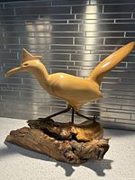 Side-angle view of the hand-carved wooden roadrunner sculpture with elongated tail and detailed beak, mounted on natural driftwood base placed on a kitchen counter with tile backsplash.