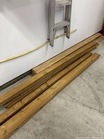 Photo shows several wood boards stacked horizontally against a white wall on concrete floor. Various thicknesses and widths visible.