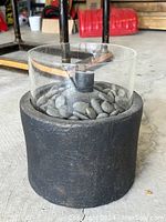 Photo of black cylindrical tabletop propane fire pit showing side view with crushed black stones and glass wind guard around burner.