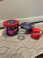 Front angle view showing all four items: two large decorative tins, small glass container, and red plastic popcorn maker.