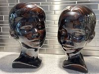 Pair of brown glazed ceramic heads representing boy and girl, placed side by side against a tiled backsplash. Good condition and glaze sheen visible.
