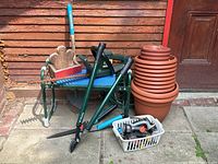 Full view of garden tools lot arranged outside against wooden shed, showing green garden bench, pots stacked, garden shears, rake, basket of hose connectors, and hedge trimmer