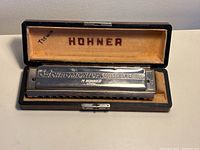 Front view of the silver metal Hohner harmonica sitting inside a black case lined with beige velvet. 'HOHNER' is printed inside the case lid.