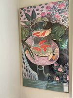 Framed poster leaning in a corner showing colorful impressionist style art with fish in a glass bowl on a pink table surrounded by plants.