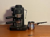 Front view of the Krups IL Primo espresso machine with glass carafe and stainless steel milk frothing pitcher with black handle, shown on wooden surface against neutral background.