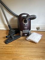 Full view of burgundy/silver Miele S4 canister vacuum with hose, floor heads, and bag of filters on wood floor