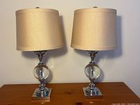 Front view of the pair of chrome and lucite table lamps with beige shades on wooden table.