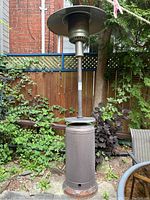 Full view of the outdoor propane patio heater placed outside, showing the tall metal structure with a circular reflector at the top and visible rust on the base.