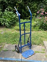 Blue two wheel dolly hand truck with black rubber wheels and metal handles with black grips, positioned upright on paved surface outdoors with garden background.