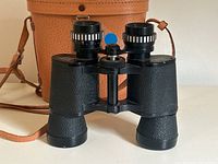 Front view of the black textured Milo binoculars showcasing adjustable focus ring and eyepieces with original leather carrying case in background