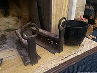 Photo showing one cast iron andiron placed front view and black metal bucket behind on a stone hearth surface next to fireplace opening