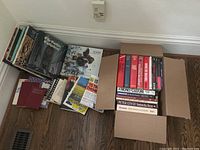 Photo showing two boxes filled with various non-fiction books laid on hardwood floor, titles visible on spines include financial and history topics