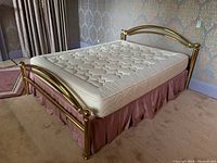 Queen size brass frame bed with Natura mattress placed on top, viewed from front angle. Shows full headboard with two vertical posts and curved top rail, mattress fully supported.