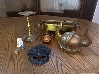 Lot of decorative brass and copper items displayed on a wooden surface including helmet, candlestick, basket, metal mask, pill box, and figurines.