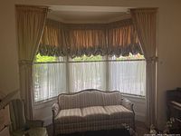 Wide shot of bay window with curtains installed, includes beige large curtain panels with tie backs, gathered valence panels on top, and sheer curtain panels below.