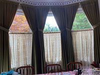 Photo showing the full bay window installation with six green velour outer panels tied back and three sheer lace panels in front, illustrating the custom fit and style of the curtains.