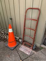 Photo showing the red metal dolly with some tire wear, positioned next to the reflective traffic pylon against a wall with rocks on the ground.