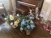 Photo showing multiple faux floral arrangements placed on the floor near a piano and furniture, displaying white, purple, pink, and blue flowers.