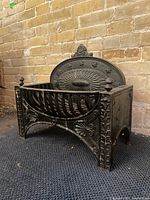 Side and front angle of black cast iron fireplace grate showing ornamental finials and embossed backplate.