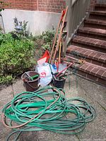 Photo showing the full lot including long-handled shovels, an axe, black buckets holding hand tools, garden hoses on cement ground, and wire trellises leaning against the step wall.