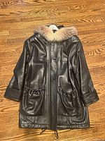 Front view of black leather jacket showing fur-trimmed hood and two large front ruched pockets with drawstrings.
