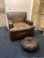 Side angle of the brown leather lounge chair and matching round ottoman on carpeted floor against brick wall.