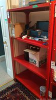 Red metal shelving unit with 3 shelves containing various boxes and items. Shelves and frame are bright red metal with bolt holes for adjusting shelf height.
