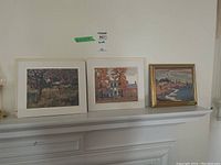 Three artworks arranged on a white mantel: two unframed prints with white matting side by side and one smaller framed painting with gold frame on the right.