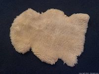 Top-down view of the natural cream sheepskin showing its full irregular shape and fluffy texture, placed on dark carpeted floor
