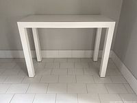 Front-facing photo showing the tabletop and legs of the white laminated rectangular table on a white tiled floor against a grey wall.