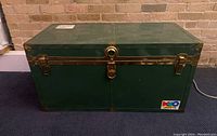 Front view of green metal trunk with metal edge reinforcements and two latch clasps, central lock, and handles on sides.