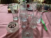 Photo showing a variety of glass vases on a pink tablecloth, including hexagonal, ribbed cylindrical, cube-shaped, and vases with unique shapes and metal adornment, plus a square glass ashtray-like piece.