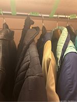 Side view of multiple winter jackets hanging on rack, including black and green items, with the blue and beige Obermeyer jacket visible.