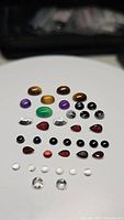 Photo showing an assortment of polished natural gemstones on white background, including tiger's eye, purple amethyst, green onyx, black and white snowflake obsidian, clear crystal quartz, white moonstone, red Mozambique garnet, and colorless white topaz.