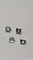 Four square cut natural aquamarine gemstones placed on a white surface, showing light blue color and clarity.