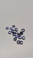 18 small round cabochon natural purple iolite gemstones on a gray surface, showcasing varying purple hues and translucency.