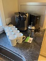 Photo showing 10 white ceramic mugs, black Keurig coffee maker, black Cuisinart coffee grinder, and Bodum French press on kitchen counter.