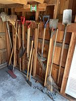 Garden shed corner with wooden rack holding multiple wooden handled gardening tools including metal shovels, garden hoes, and rakes.