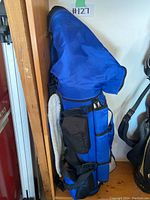 Blue golf bag leaning against wall with multiple side and front pockets, padded with shoulder strap visible.