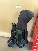 Two golf bags side by side against beige wall; one black Callaway stand bag, one green and black cart bag