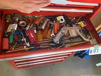 Top-down view of a red metal toolbox drawer filled with assorted hand tools and hardware including screwdrivers, scissors, and utility knives.