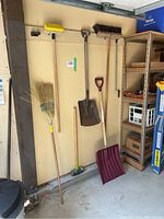 Various yard tools including two snow shovels, two brooms, and one floor brush hung on a garage wall.