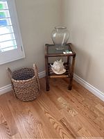 Photo showing bamboo side table with glass top and a clear glass vase on top, conch shell on lower shelf, and woven basket next to table on hardwood floor by window.