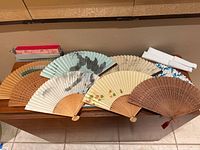 Top-down view of 7 different Asian folding fans including wooden and fabric fans showing various colors and patterns