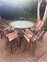Six piece patio set on brick patio including one round table with glass top and stone inlaid metal base, with five high back metal chairs with striped cushions.