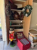 Full view of open cabinet showing several Christmas wreaths decorated with seasonal items, a large classic nutcracker figure standing beside, and red storage bins with organized ornaments