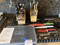 Photo of two wooden knife blocks holding multiple kitchen knives with various handle types, plastic case with Trusted Butcher knife set inside, and additional knives with guards laid out on stove