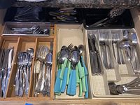 Various pieces of flatware including forks, knives, spoons with blue and green plastic handles and stainless steel utensils organized in trays