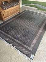 Full view of rectangular hand woven floor rug placed near carved wooden furniture, showing overall pattern and fringe detail.