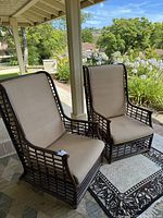 Two large high backed bamboo chairs with beige cushions placed outdoors on a covered patio. Chairs positioned side by side on a patterned rug, showing the lattice bamboo frame and high backrest design.